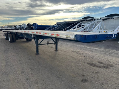Flatbed trailer shown in side view with blue frame and long rectangular deck, multiple axles, and support legs extended. Text on side rail includes reflective tape and small labeling.