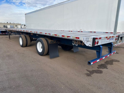Flatbed trailer in side view showing dual rear axles, white wheels, and reflective safety tape along sides.