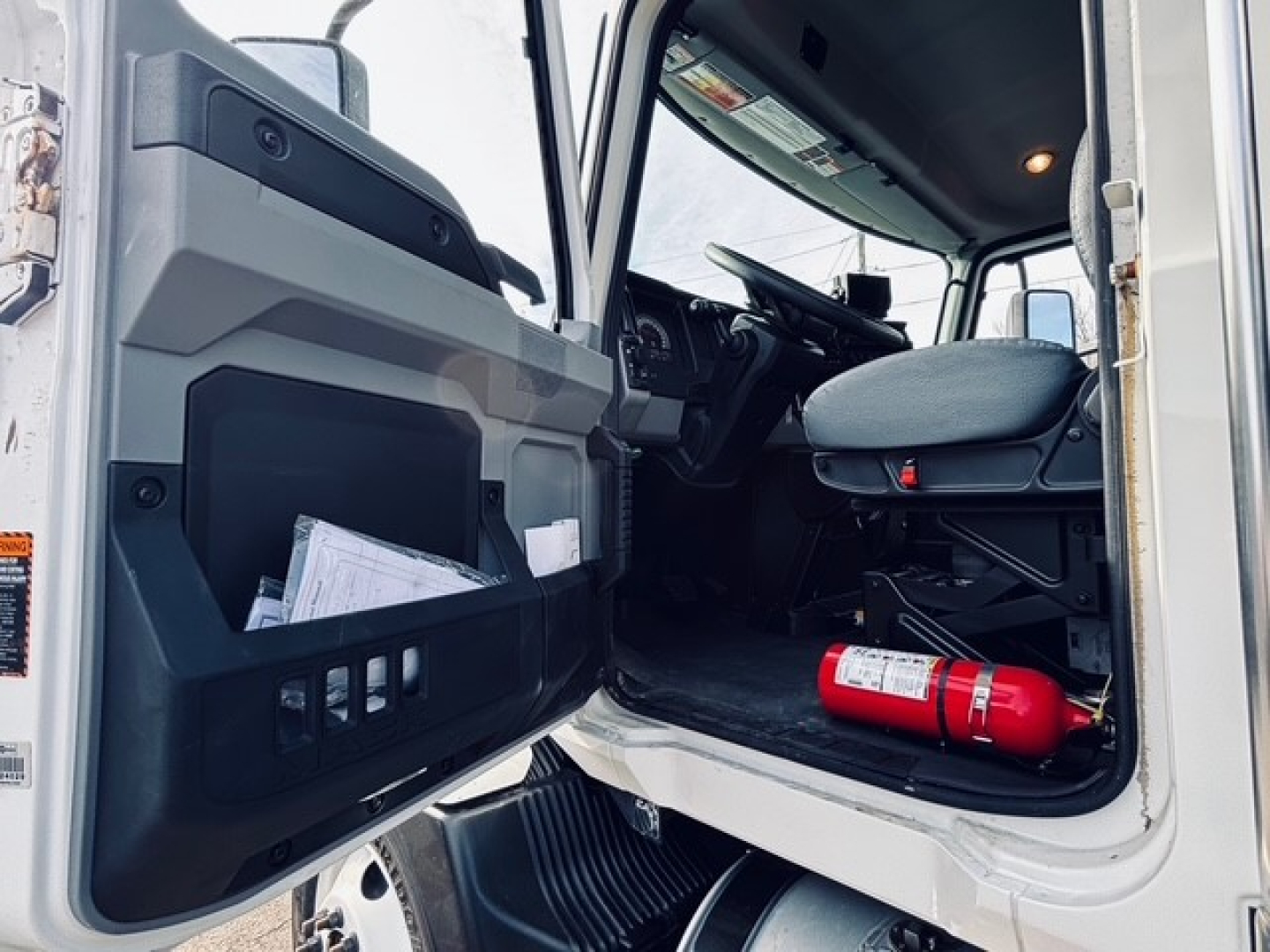 Cab interior of a 2025 Mack MD6 dump truck showing open door, steering wheel, driver's seat, and a red fire extinguisher on the floor. Visible door storage pocket and steps.