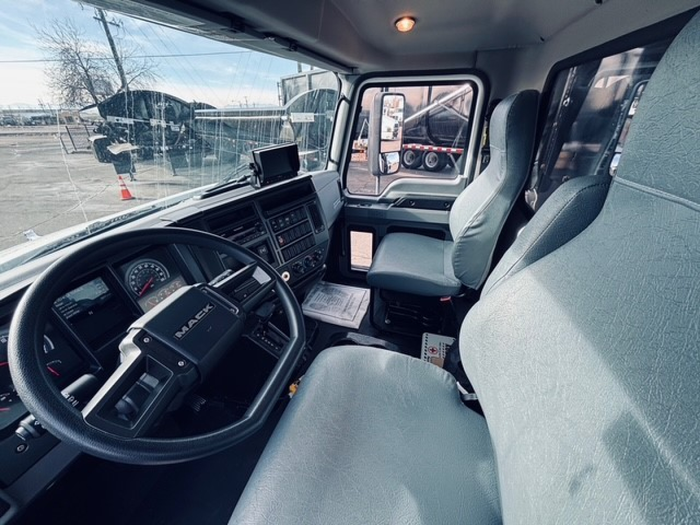 Cab interior of a 2025 Mack MD6 dump truck showing steering wheel, dashboard, driver seat, and passenger seat with light fabric upholstery and side window.