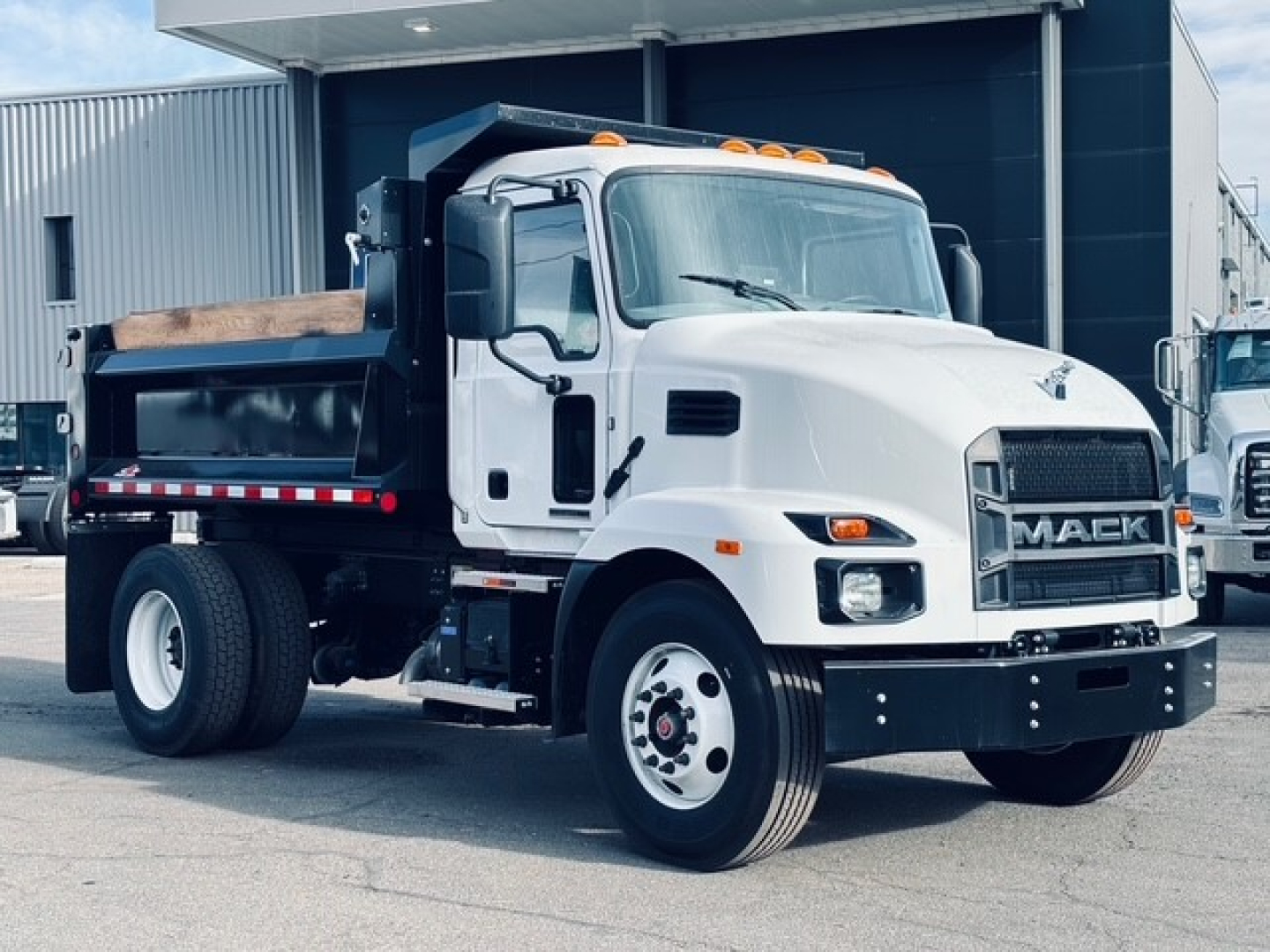 White 2025 Mack MD6 dump truck in a dealership lot showing front view and dump bed with black grille and side mirrors.