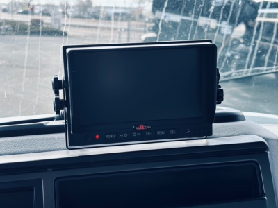 Cab interior showing a dash-mounted monitor/screen. The screen is centered on the dash with small control buttons below. The windshield outside shows a blurred exterior.