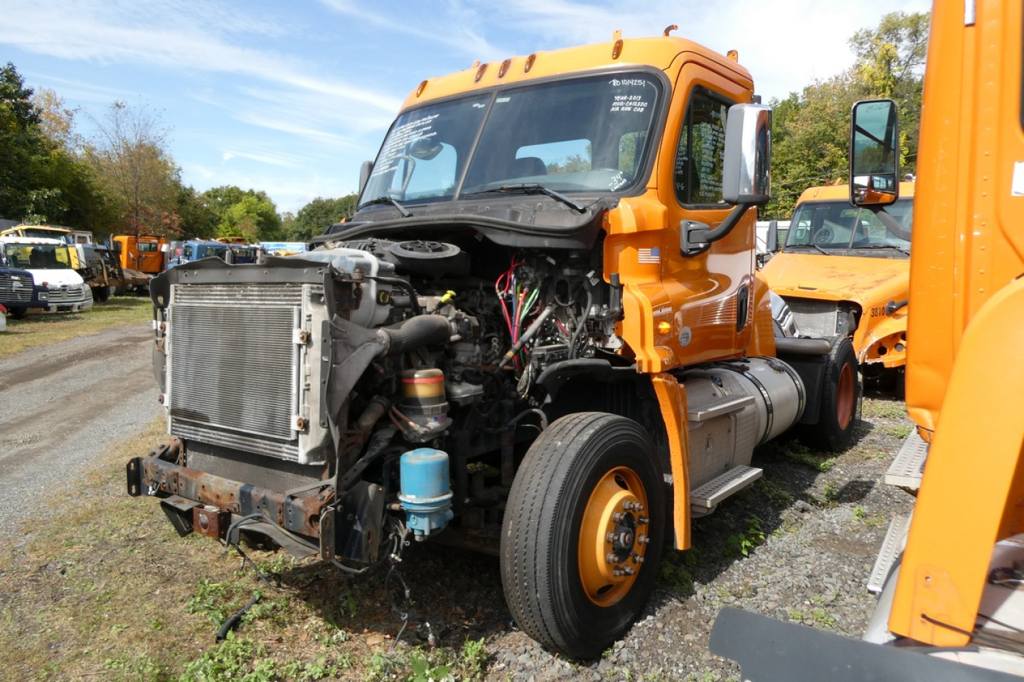 Front view of a 2013 Freightliner Cascadia 125 daycab truck with exposed engine bay and radiator, orange cab, and other orange trucks in a yard.