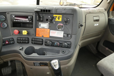 Dashboard of a Freightliner Cascadia 125 cab showing steering column, controls, and gear shifter. Visible text includes '6-2' yellow sticker, warning labels, and various knobs on the center panel.