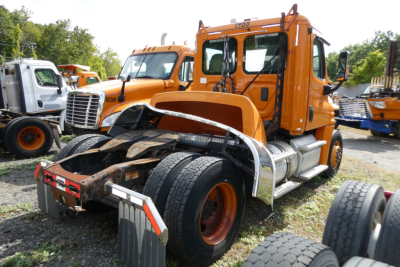 Orange Freightliner Cascadia 125 daycab truck in a lot with other orange trucks; front view shows damaged bumper and exposed frame with dual rear wheels.