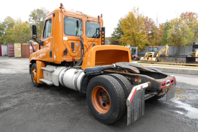 Truck cab and rear chassis of a 2013 Freightliner Cascadia 125 daycab showing orange exterior, fuel tanks, and rear wheel with mudguard.