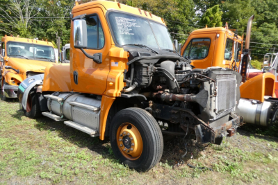 Orange Freightliner Cascadia 125 cab tractor with exposed engine bay and front end; missing bumper, visible engine components, single front wheel; other orange trucks in background.