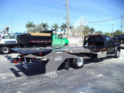 Ram 5500 rollback tow truck with flatbed in parking lot, side view showing chain and hook, diagnostic stickers on cab, palm trees in background