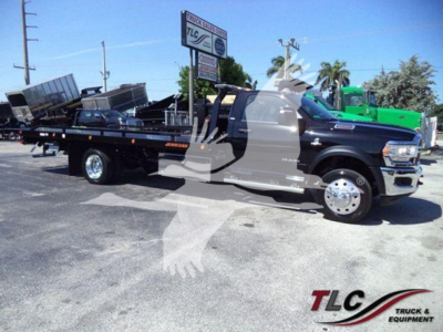 Ram 5500 rollback tow truck side view showing flatbed with equipment and Ram badge, blue cab, black bed, dealership lot in background.