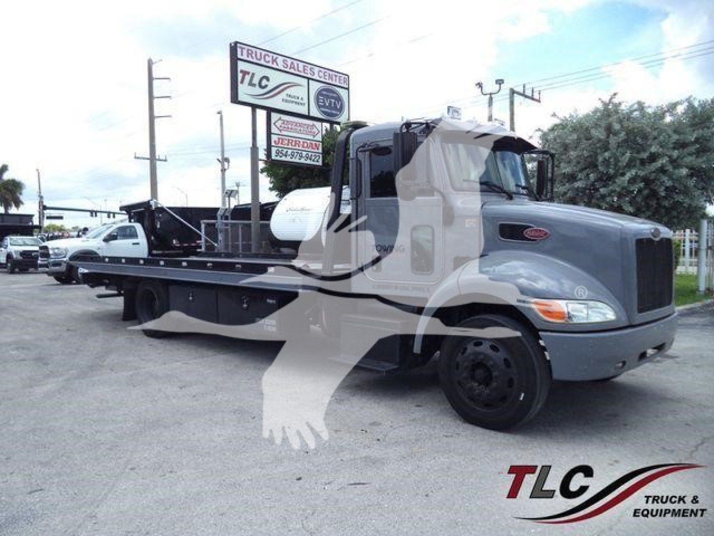 Peterbilt 337 roll-back truck with extended flatbed showing cab and front wheel, dealership sign in background. Visible text on truck and sign includes 'Peterbilt', 'TLC Truck Sales Center' on the sign, and numbers on the sign.
