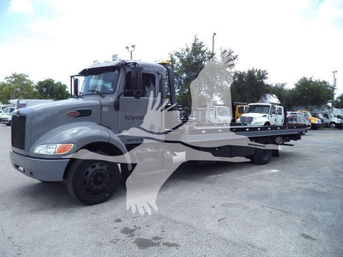 Peterbilt 337 rollback tow truck in a used inventory lot, gray cab with flatbed, side mirrors, and multiple trucks in background.