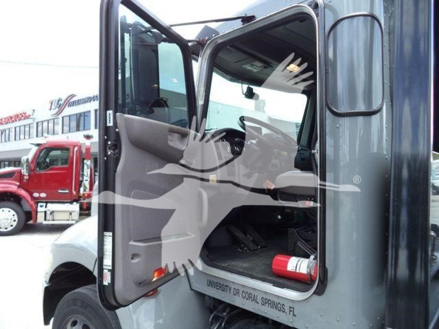 Truck cab interior with open door showing steering wheel and pedals, side mirror, and a fire extinguisher strapped inside; text on dashboard area and door trim not fully legible.