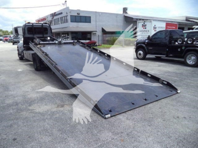Peterbilt 337 rollback tow truck with long black metal ramp extended in front, service shop background, VIN plate not visible.