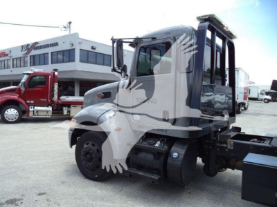 Peterbilt 337 rollback truck side view showing cab, fuel tank, and chassis with wheel; background shows dealership building and another red truck.