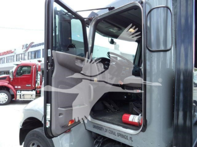 Truck cab interior with open door showing steering wheel and pedals, side mirror, and a fire extinguisher strapped inside; text on dashboard area and door trim not fully legible.