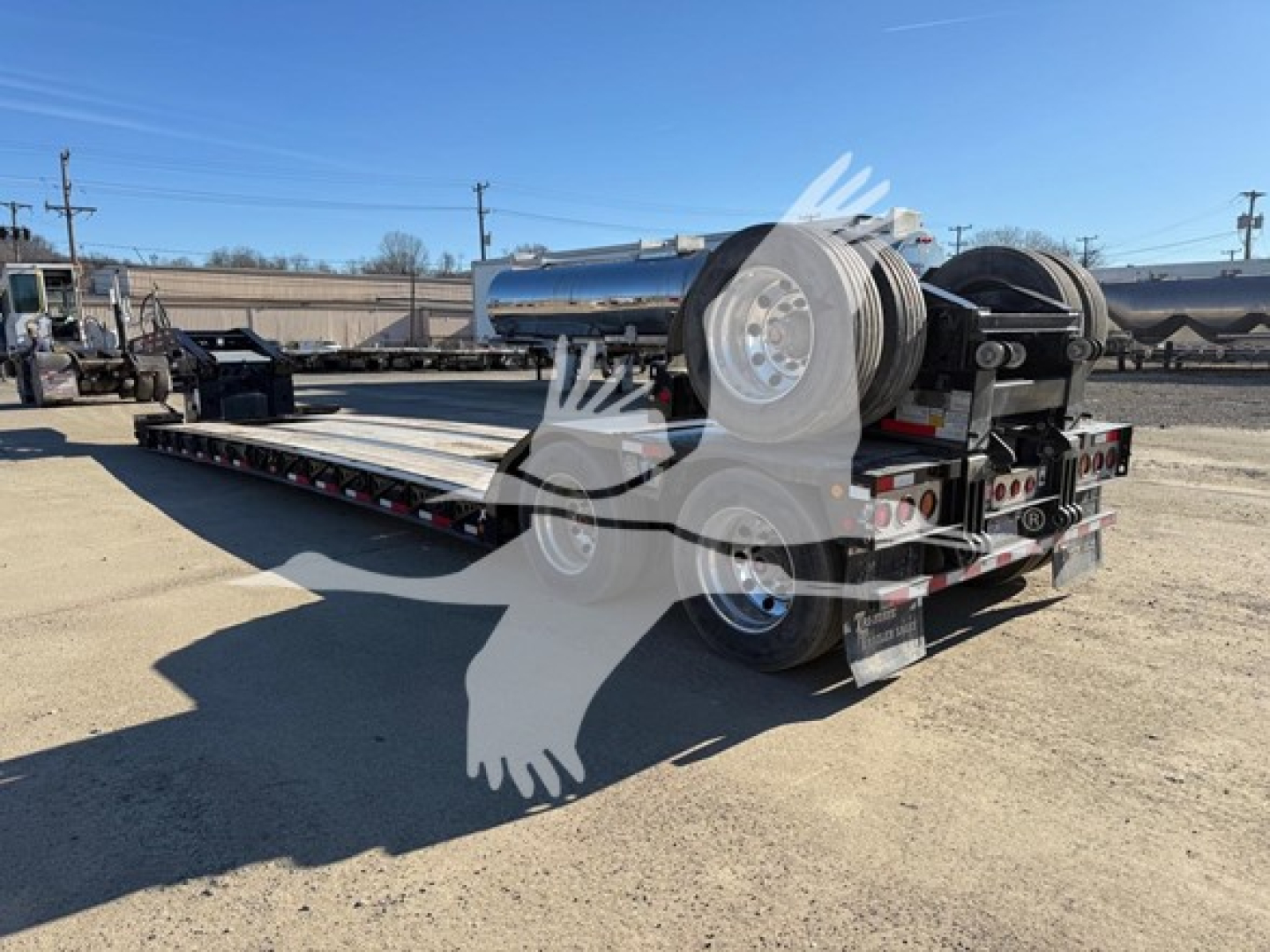 Lowboy trailer with dual rear axles and chrome fuel/tank units, viewed from side at an outdoor lot; long flatbed deck, multiple wheels, and a rear winch/axle assembly visible.
