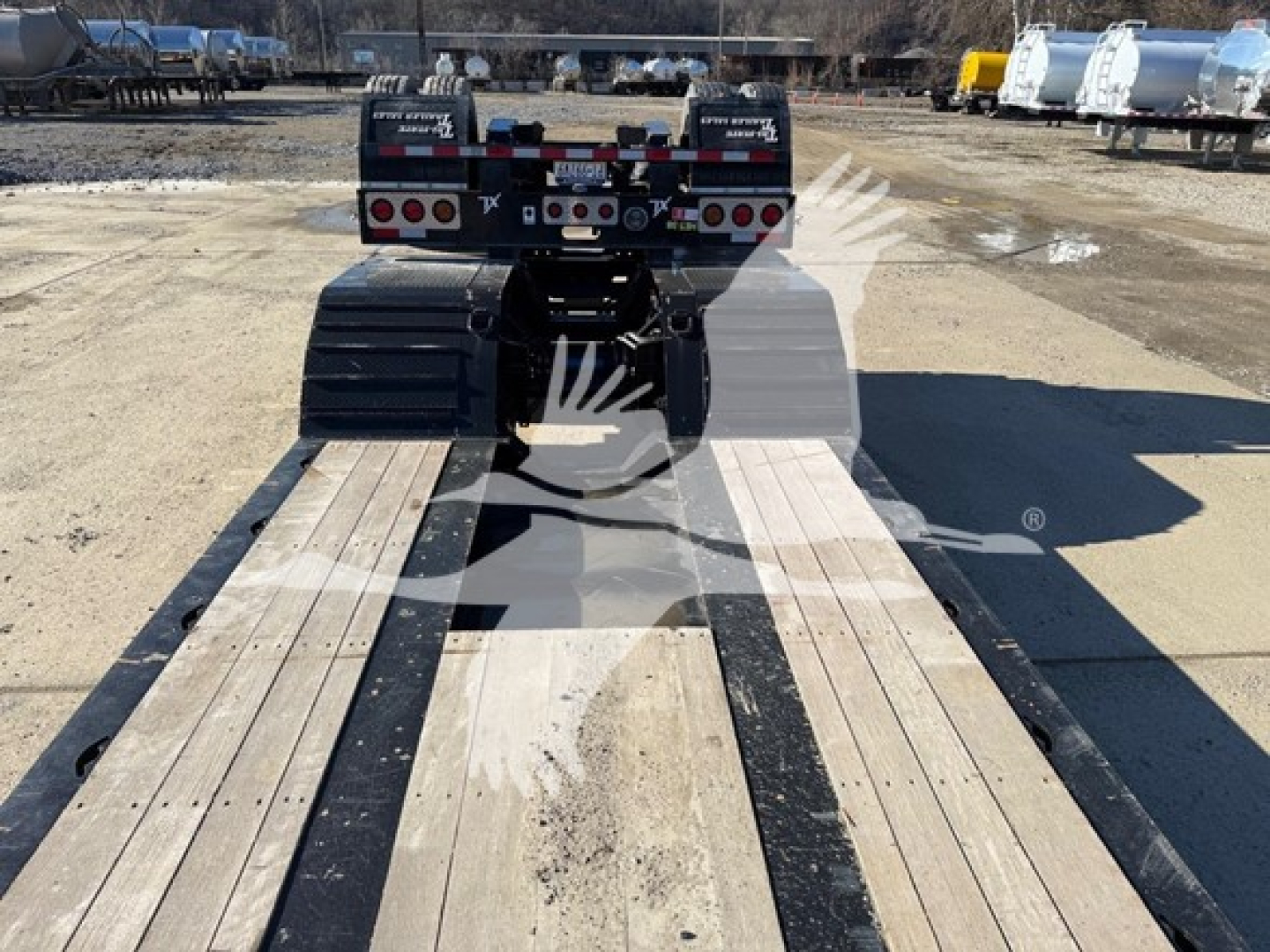 Lowboy trailer in a yard showing a gooseneck hitch and dual ramps with deck planks, rear view with tires and LED lights.