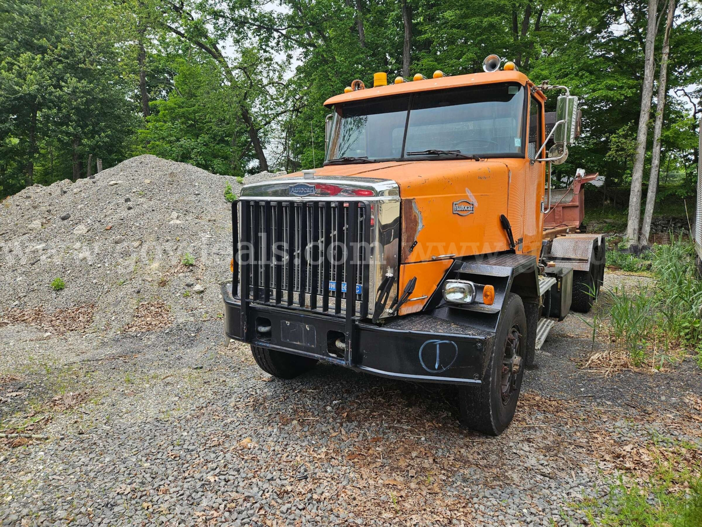 Orange Autocar roll-off truck in a gravel lot with pile of dirt in background. Front grille with chrome vertical bars, large bumper, two round headlights, amber beacon lights on cab roof. Some text on side panels and cab.