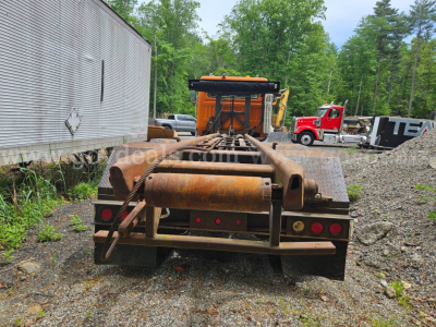 Rear view of a used roll-off truck trailer with rust-colored metal frame and a hydraulic winch, ATV-style back lights, gravel ground, trees in background.