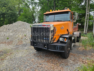 Orange Autocar roll-off truck in a gravel lot with pile of dirt in background. Front grille with chrome vertical bars, large bumper, two round headlights, amber beacon lights on cab roof. Some text on side panels and cab.