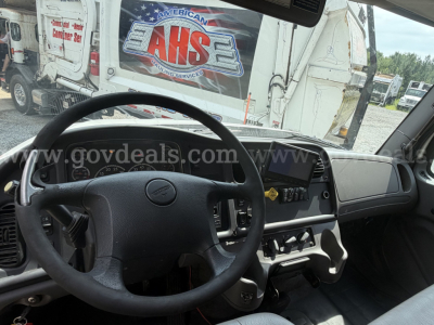 Truck cab interior of a 2015 Freightliner 114SD showing steering wheel, dashboard, and center console with display screen; background shows a recycling/garbage truck and branding.