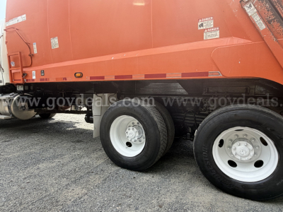 Truck side of a 2015 Freightliner 114SD dump truck showing rear wheels and black undercarriage with warning labels on orange dump body.