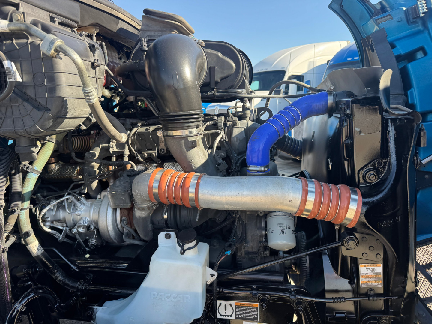 Peterbilt 579 engine bay showing large turbo intake and hoses, blue silicone hose, air intake piping, orange-coupled intercooler pipe, coolant reservoir, and various labels.