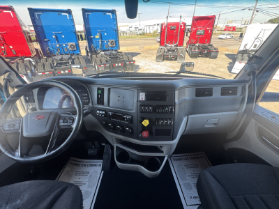 Peterbilt 579 truck interior dashboard and steering wheel with control panels and storage, view from driver seat; multiple colorful trucks visible outside.