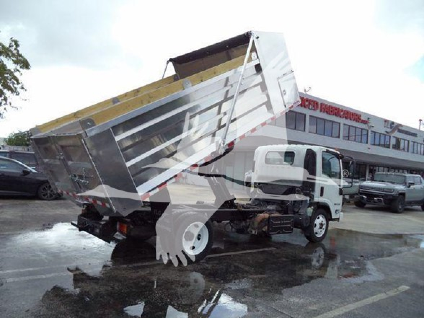 Isuzu NPR XD dump truck with tilted metal dump bed raised, rear view showing suspension and wheels; dealership lot in background.