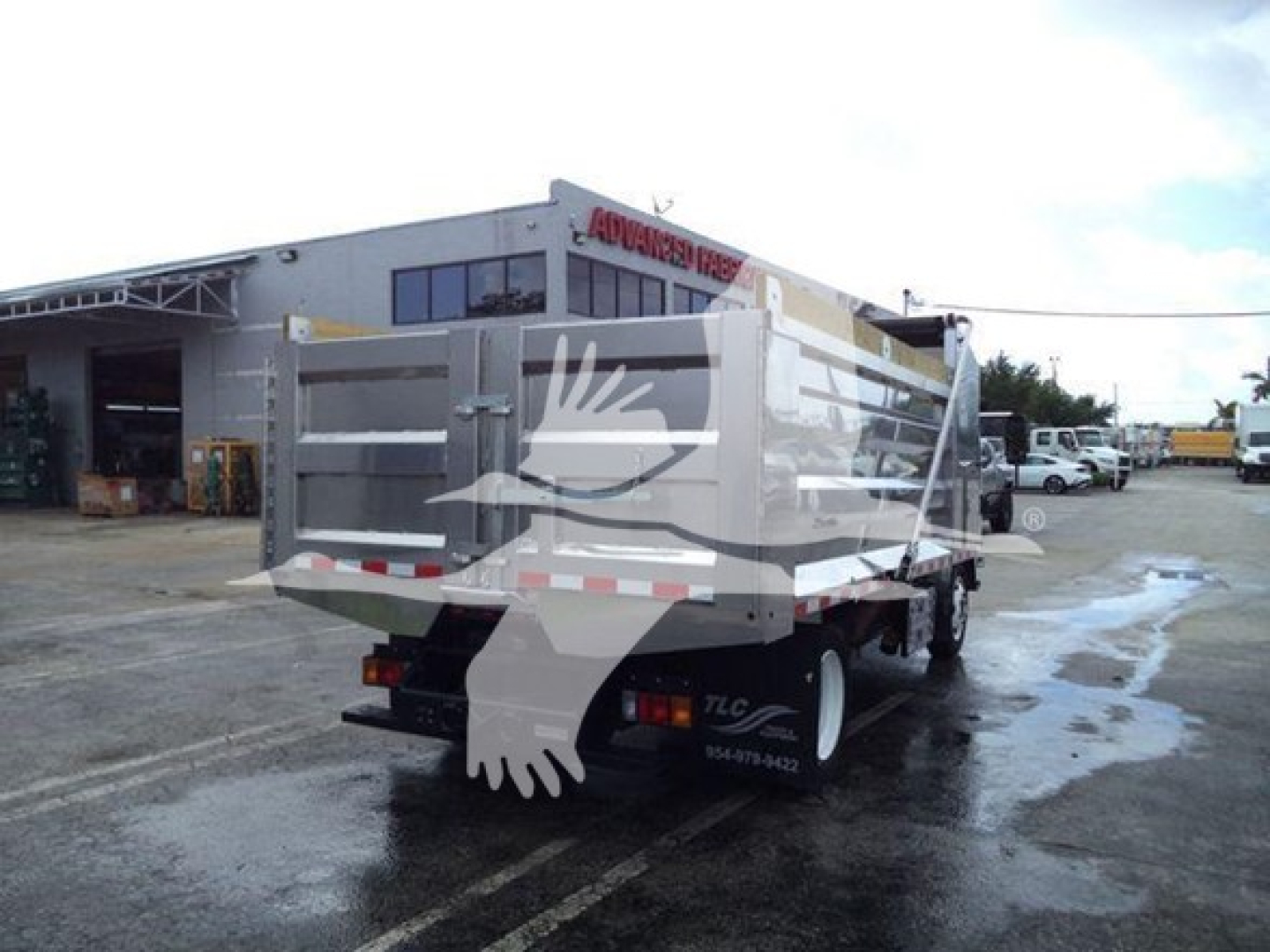 Isuzu NPR XD dump truck rear view showing dump bed and taillights; dealership background with building and lot.