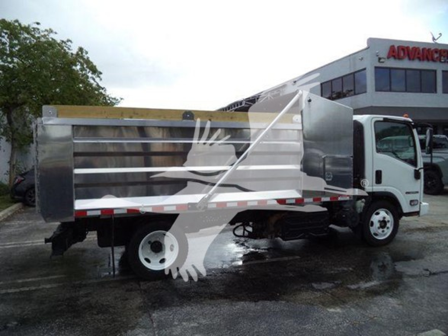 Isuzu NPR XD dump truck side view showing aluminum dump bed and cab, with a white truck and dealership building in background.