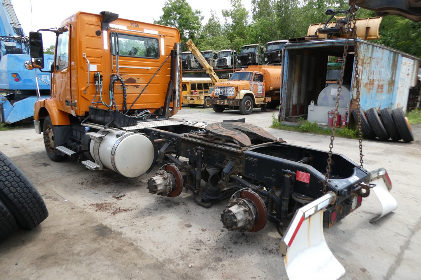 Orange Volvo truck cab with exposed chassis and rear axle assembly in lot; VIN plate not visible; other trucks and tires in background.