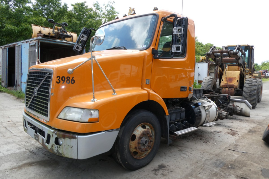 Volvo VNM64T conventional daycab truck showing front grille and orange cab with stock number 0612232 and 3986 on hood.