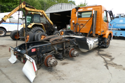 Volvo VNM64T truck chassis with exposed drivetrain and orange cab, front view showing wheels, axles, and suspension components.