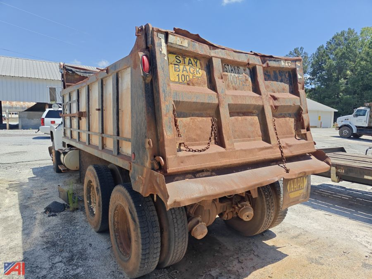 Dump truck bed with rear view showing rusted metal sides and dual rear axles; warning label Stay Back 100 and hinges on tailgate visible.
