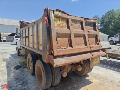 Dump truck bed with rear view showing rusted metal sides and dual rear axles; warning label Stay Back 100 and hinges on tailgate visible.