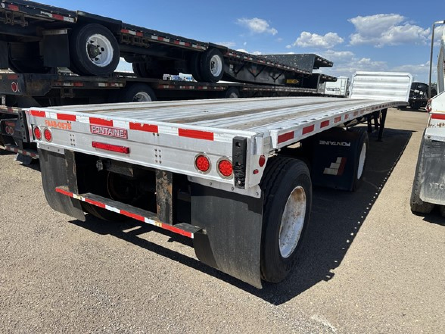 Flatbed trailer with a silver aluminum deck and red/white reflective stripes; rear view showing taillights, mudflaps, and dual axles.
