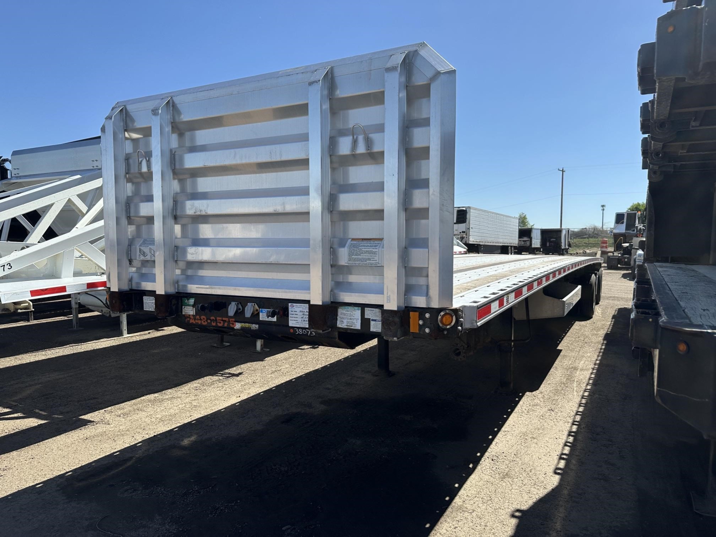 Flatbed trailer with silver steel sides and rear ramp, viewed from left rear corner, showing reflective safety lights and hinged gate.
