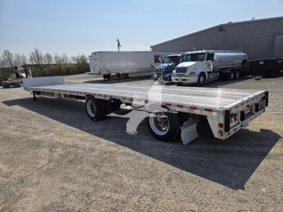 Flatbed trailer showing a double-axle rear with silver deck and side rails, rear lights, and white cab truck in the background.