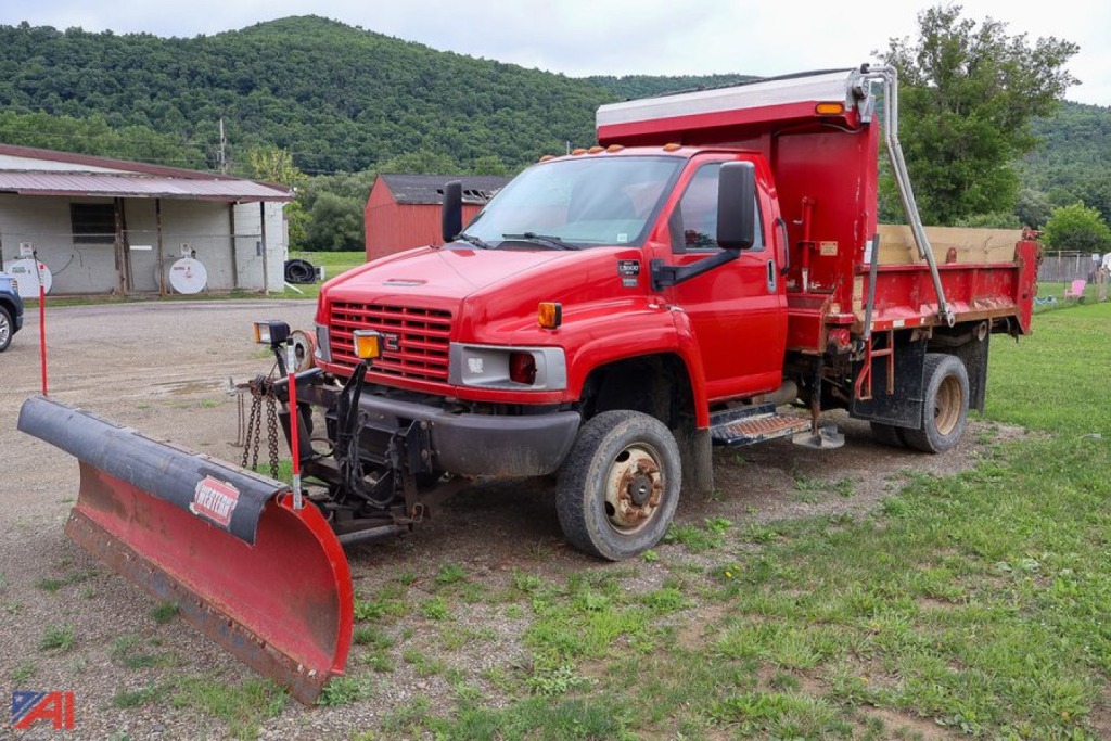 GMC TopKick C5500 dump truck with a front plow attachment, red cab and dump bed, off-road yard setting.