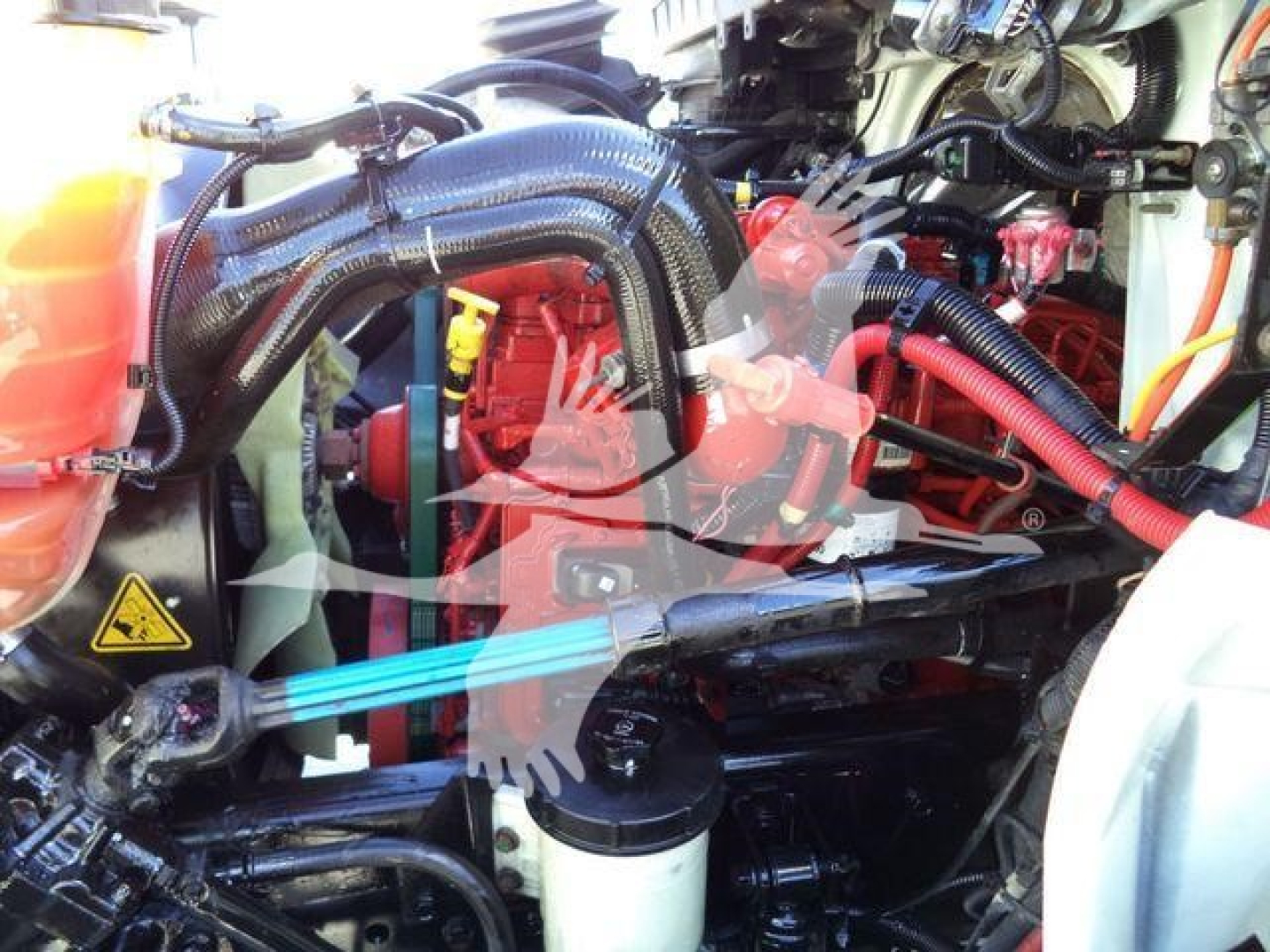 Truck engine bay with various hoses and components. Visible text includes warning triangle sticker and some component labels; no clearly legible model text.