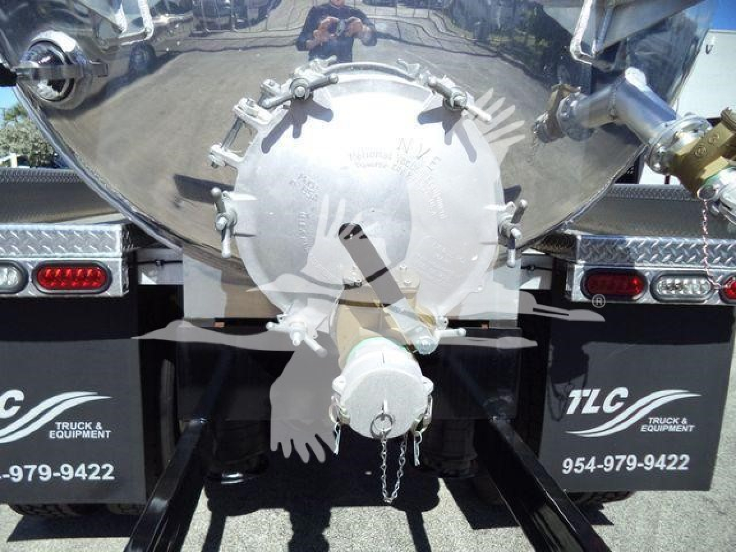 Circular rear valve assembly on a vacuum tank trailer with locking bolts and a small chain. Reflective surface shows vehicle surroundings.