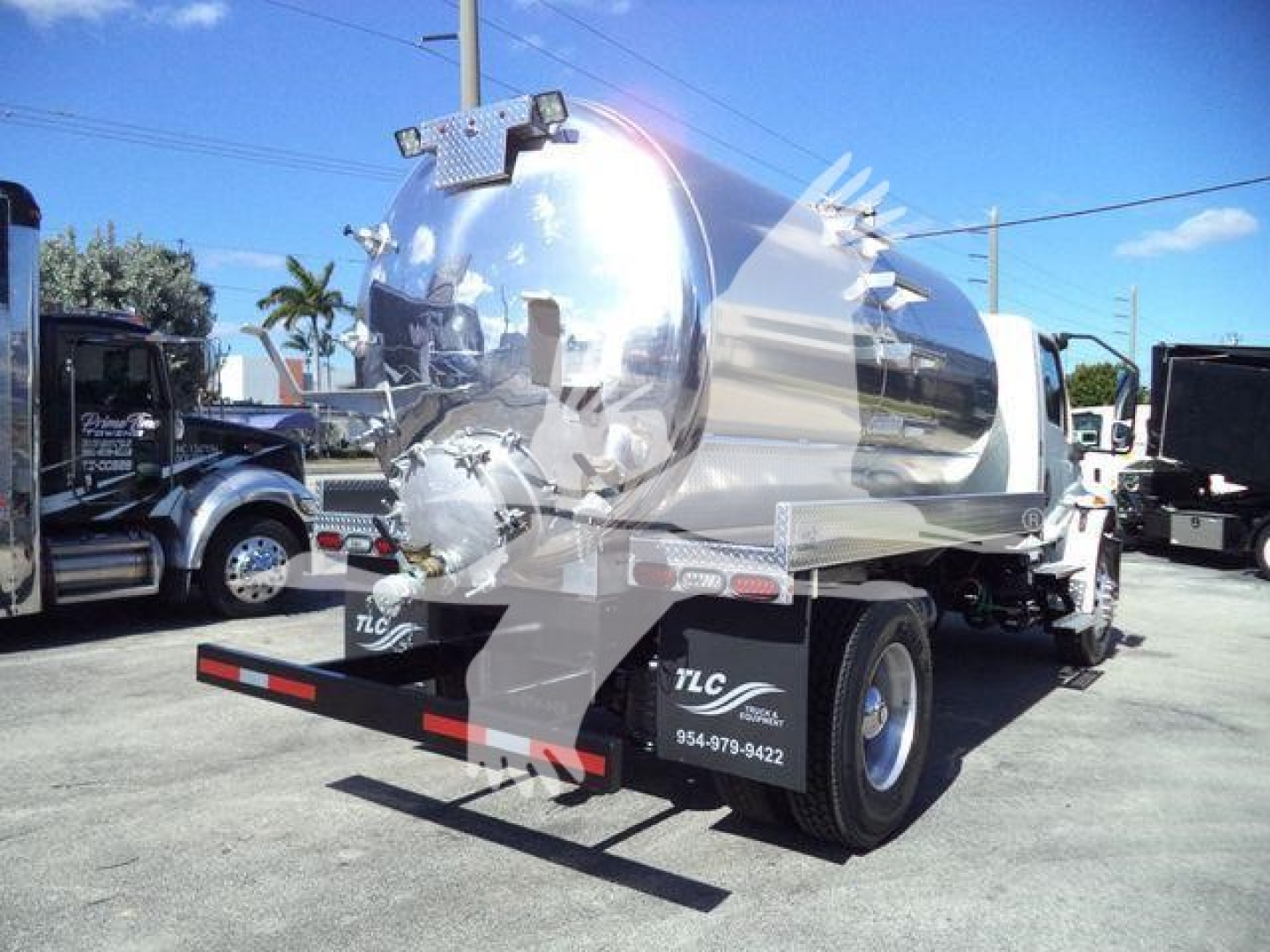Vacuum tank truck rear/side view showing polished stainless tank, dual rear axles, and black bumper. Visible text on wheel area: TLC, phone number 954-979-9422. Nozzle fittings and manhole cover on tank.