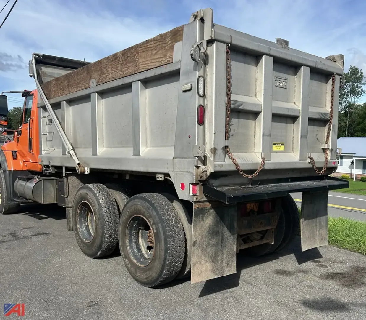 Dump truck rear showing three rear axles, tailgate with chains, metal dump bed, and wooden rail on top edge. Visible text includes nothing legible.