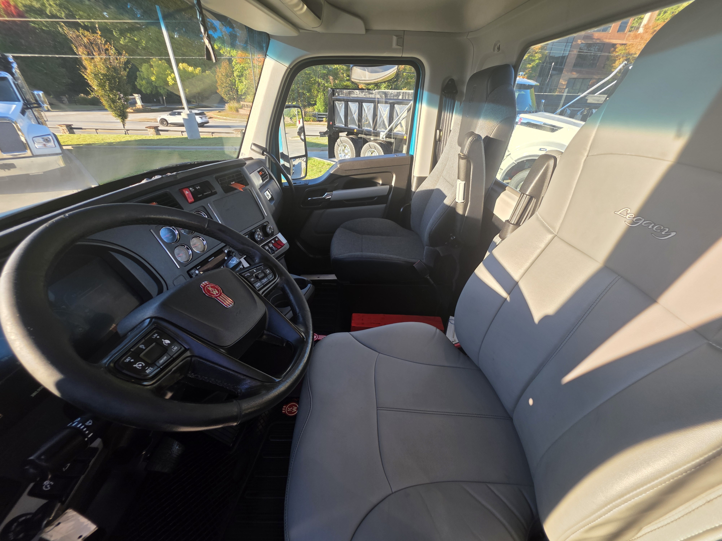 Kenworth T880 truck cab interior with steering wheel, dashboard, and driver seat visible; gray cloth seats; dashboard instrumentation; door and window; exterior side mirror reflected.