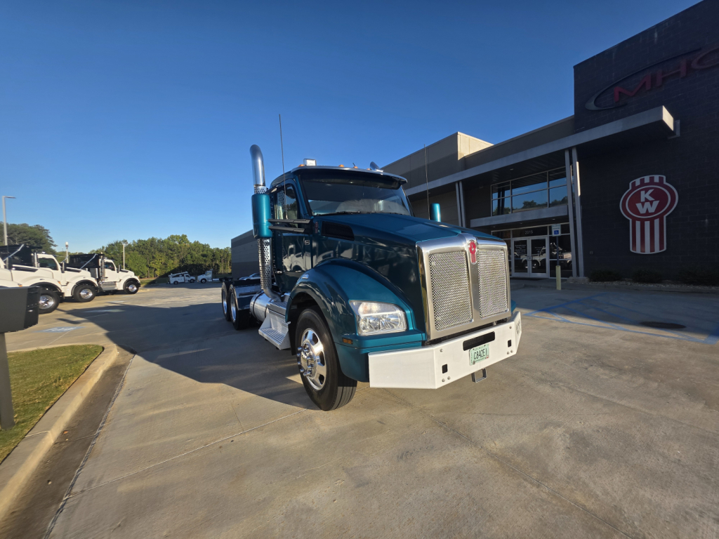 Kenworth T880 conventional daycab truck in teal/blue shown from front-left angle in dealership lot with chrome grille and large exhaust stacks.