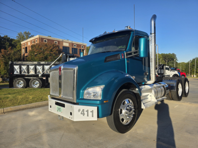 Kenworth T880 conventional daycab truck in teal/blue, front-left view showing chrome grille, large exhaust stacks, aluminum steps, and triple rear axles; white tag with number 411 on bumper; license plate area visible.