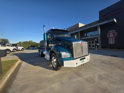 Kenworth T880 conventional daycab truck in teal/blue shown from front-left angle in dealership lot with chrome grille and large exhaust stacks.