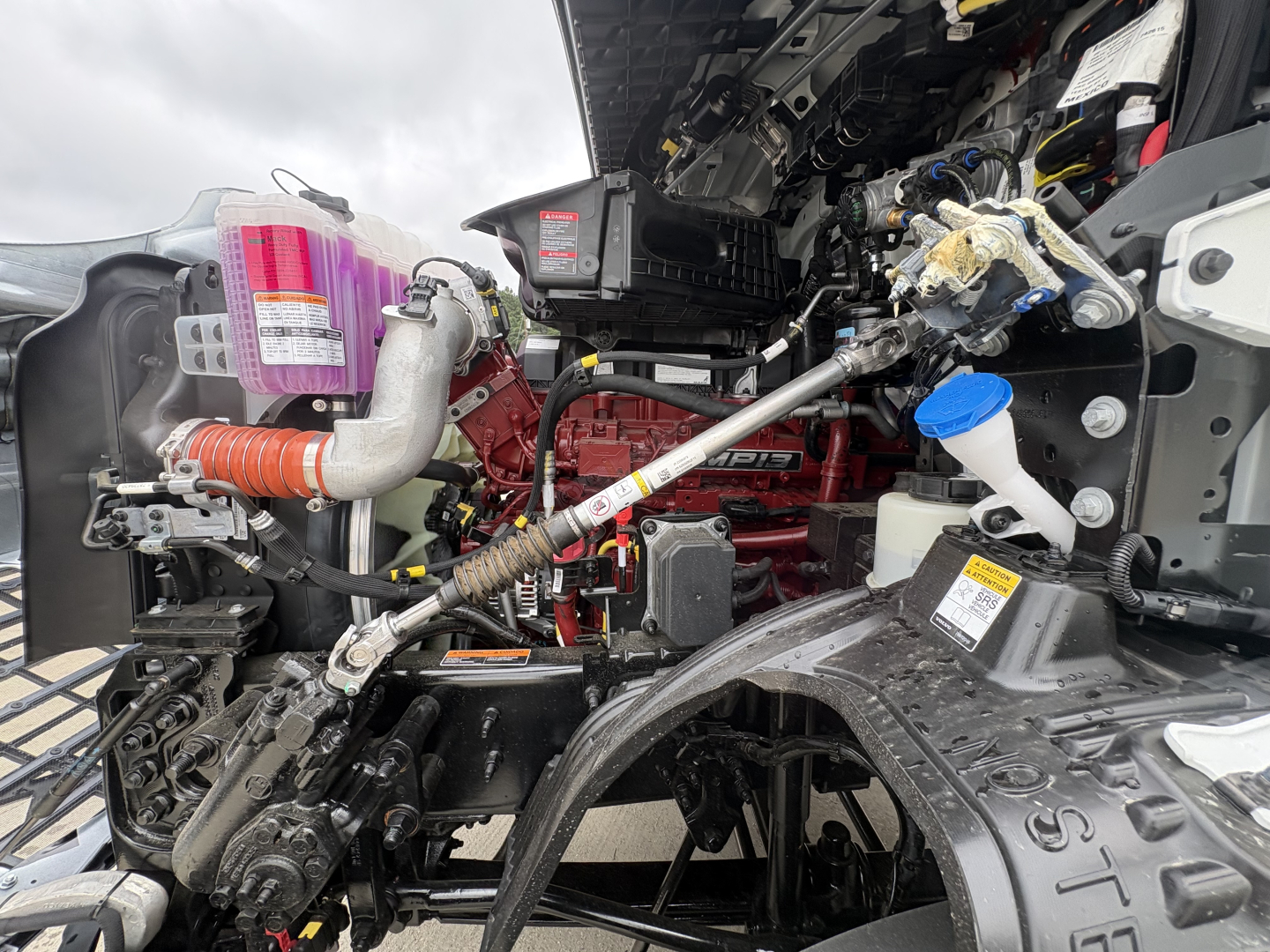 Mack AN64T engine bay showing red engine block, blue coolant reservoir cap, air intake hoses, and hydraulic lines in a new conventional daycab truck.