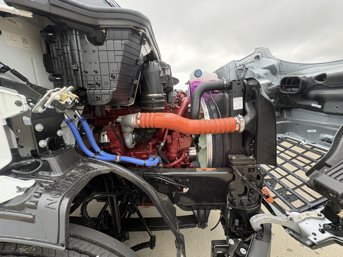 Truck engine bay showing Mack AN64T diesel engine with red blocks, orange coolant hose, blue fuel lines, radiator and intake components visible.